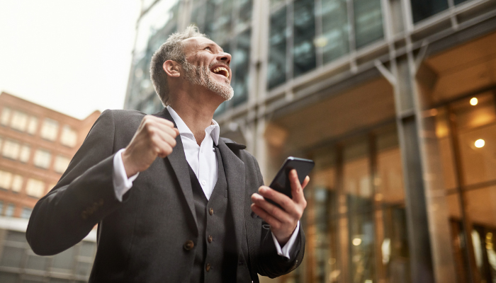 Businessman standing in front of an office building punching the air and holding his phone to denote he is happy with the winner of the comparison of NetSuite and Sage Intacct.