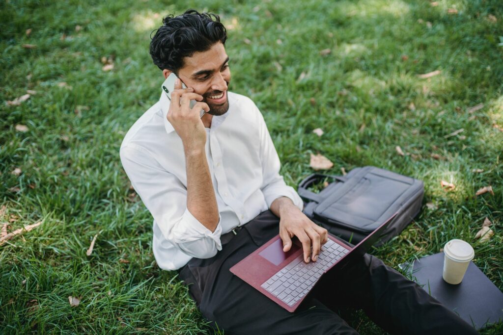 Business man sat on grass with his laptop and smiling while on his phone, denoting access to NetSuite support from anywhere.